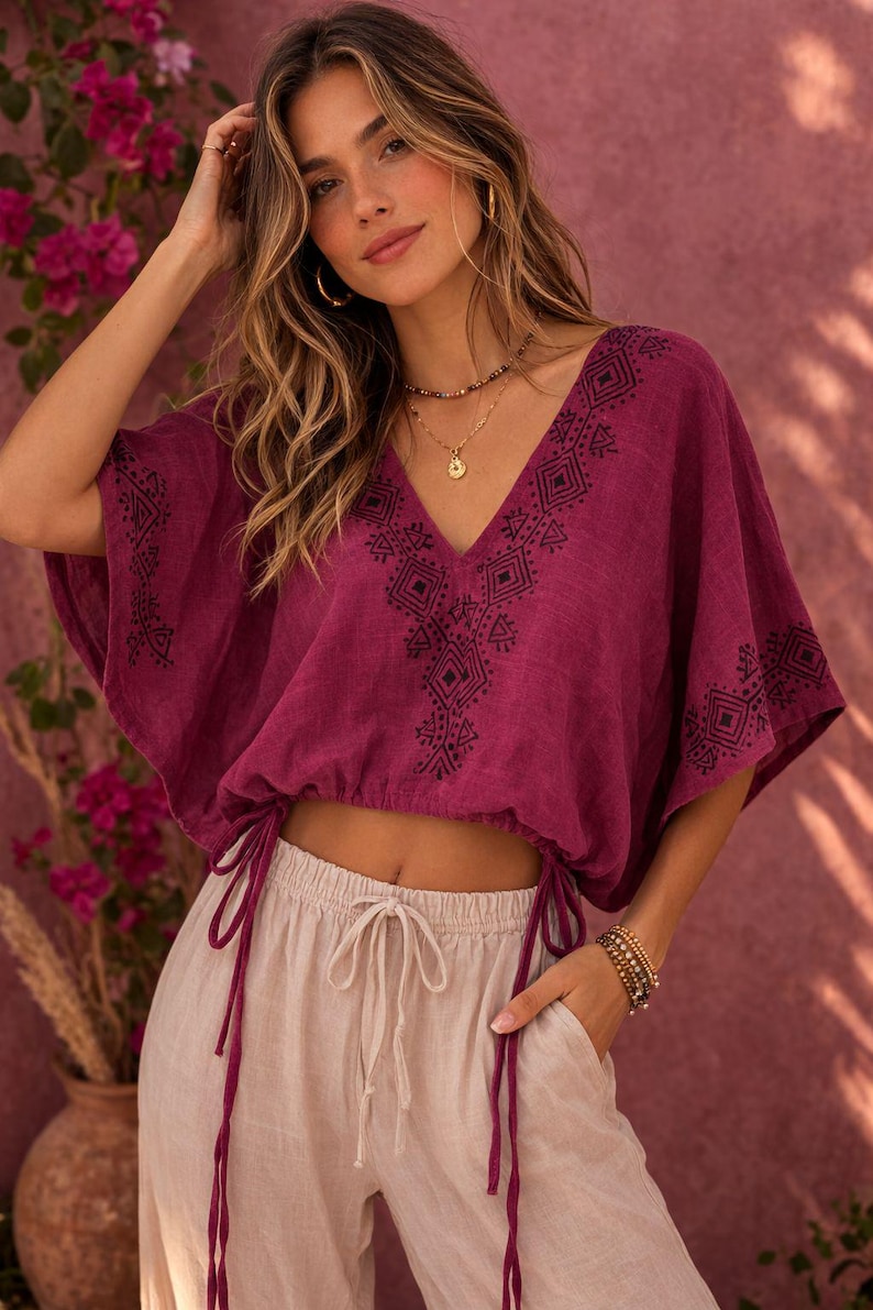 May include: A deep magenta cropped blouse featuring a V-neckline and wide short sleeves, both adorned with black geometric embroidery. The hem includes an adjustable drawstring with long ties on each side, paired with light beige drawstring pants.
