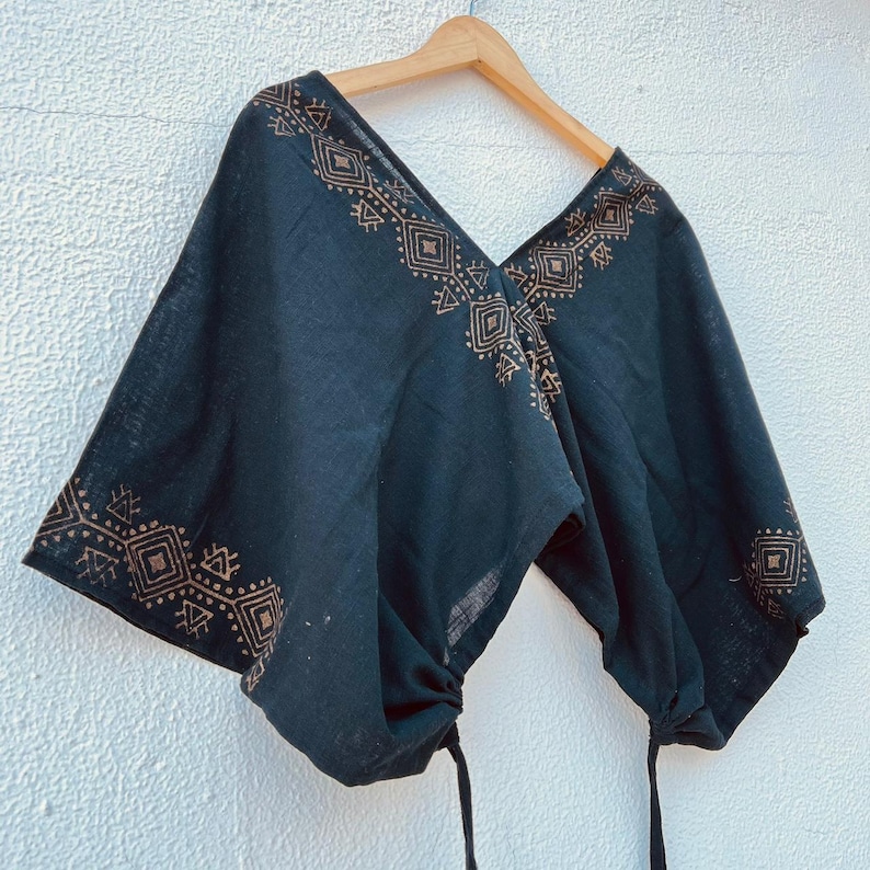 May include: Black top with a V-neck and short sleeves. The top features a geometric pattern in a lighter brown color along the neckline and sleeve edges. The sleeves are gathered with drawstrings. The top is hanging on a wooden hanger.
