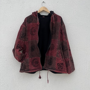 May include: A red and brown patchwork hoodie with a black interior, featuring a full zip and a hood. The jacket is decorated with celestial and floral patterns. The jacket is hanging on a wooden hanger.