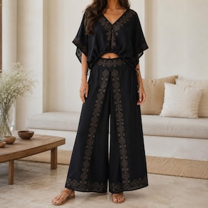 May include: Black two-piece outfit featuring a V-neck top and wide-leg pants. The top has short sleeves and drawstring details. The pants have a decorative pattern down the front. The outfit is made of a lightweight fabric.