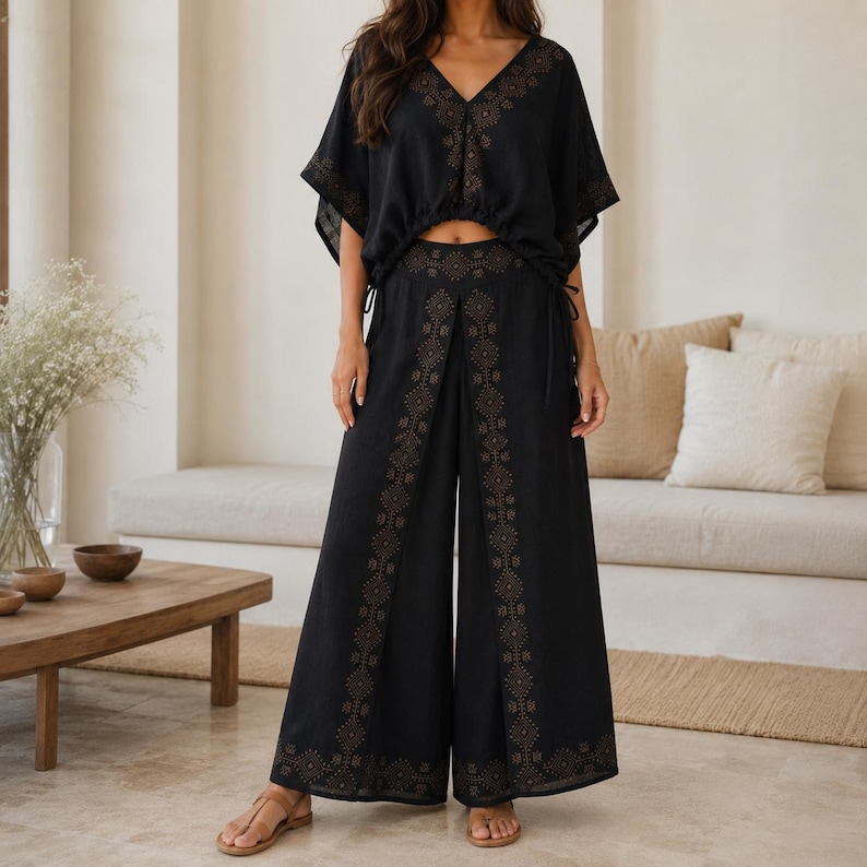May include: Black two-piece outfit featuring a V-neck top and wide-leg pants. The top has short sleeves and drawstring details. The pants have a decorative pattern down the front. The outfit is made of a lightweight fabric.
