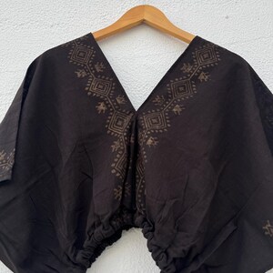 May include: A black short-sleeved top featuring a deep V-neckline and a gathered elasticized hem. The garment is decorated with a brown geometric block-print pattern that outlines the neckline and extends down the front in a diamond motif.