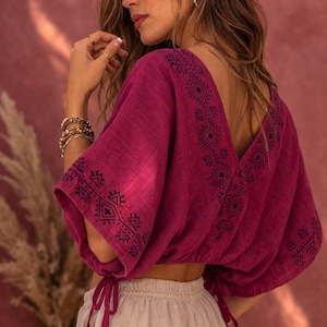 May include: A magenta cropped blouse featuring a deep V-neckline and wide sleeves with drawstring ties at the cuffs. The garment is adorned with dark blue geometric embroidery along the neckline and sleeve edges, paired with light beige trousers.