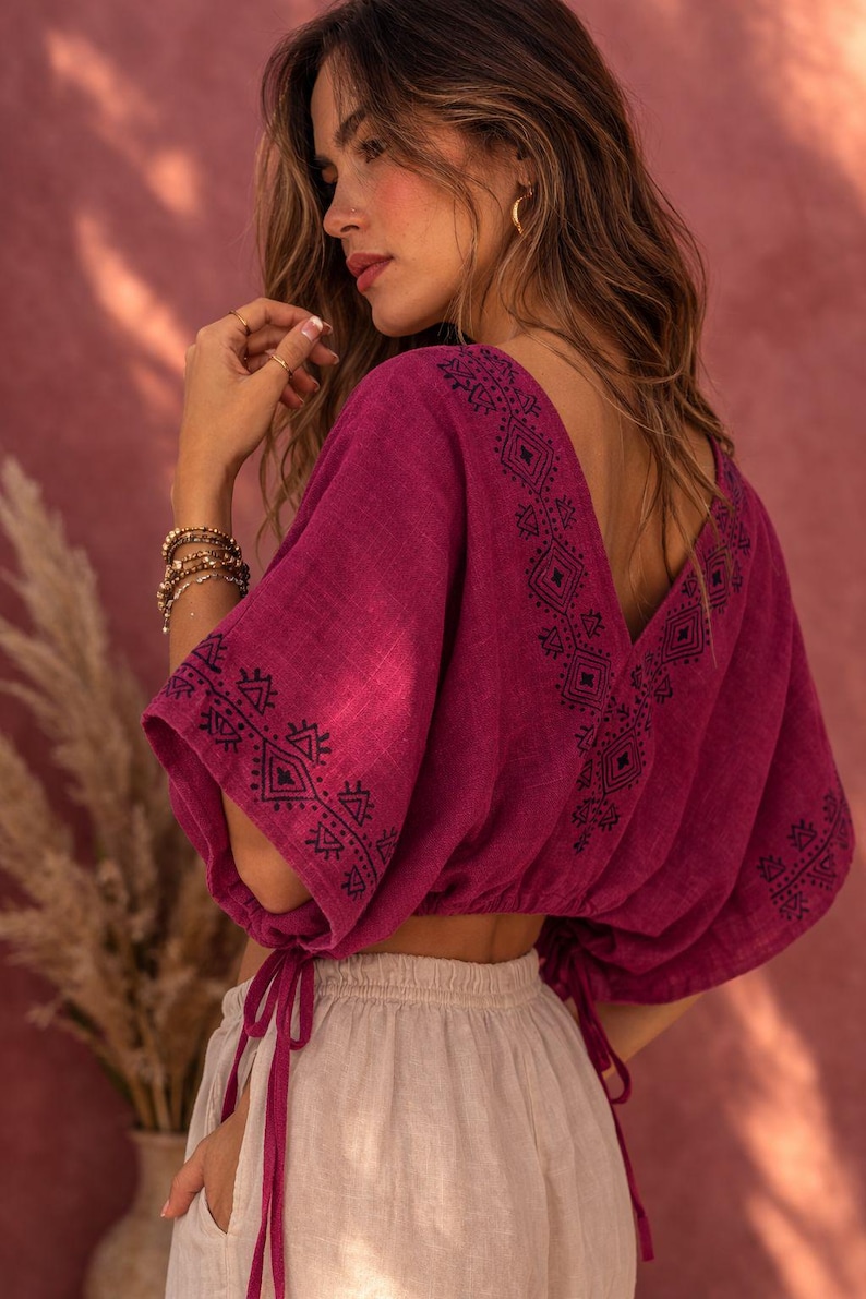 May include: A magenta cropped blouse featuring a deep V-neckline and wide sleeves with drawstring ties at the cuffs. The garment is adorned with dark blue geometric embroidery along the neckline and sleeve edges, paired with light beige trousers.