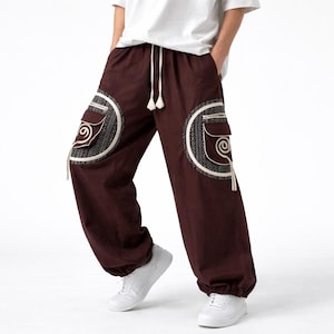 May include: Brown cargo pants with decorative circular patches on the pockets. The patches feature a cream-colored spiral design and a patterned border. The pants have a drawstring waist and elastic cuffs, and are worn with white sneakers.