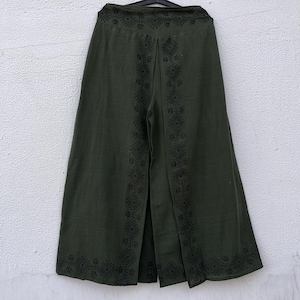 May include: Olive green wide-leg pants with a split front and decorative black geometric embroidery. The pants are hanging on a black hanger against a white wall. The embroidery is along the seams and the waistband.