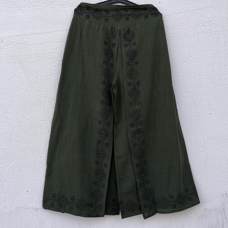 May include: Olive green wide-leg pants with a split front and decorative black geometric embroidery. The pants are hanging on a black hanger against a white wall. The embroidery is along the seams and the waistband.