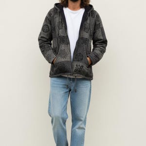 May include: A grey patchwork hoodie with a black hood and floral patterns. The jacket is open, revealing a white t-shirt. The person is wearing light blue jeans.