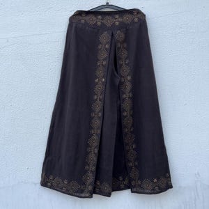 May include: Dark brown wide-leg pants with a decorative geometric pattern in a lighter brown color. The pants are hanging on a black hanger against a white wall. The decorative pattern is along the sides and top of the pants.