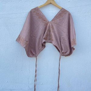 May include: A mauve-colored top with a V-neck and batwing sleeves, adorned with geometric patterns in a darker shade. The top has drawstrings at the bottom and is hanging on a wooden hanger against a white wall. The top is made of a soft fabric.