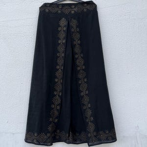 May include: Black skirt with a split front and intricate geometric embroidery in a gold color. The skirt has a high waist and a flowing, A-line silhouette. The embroidery is concentrated along the edges and the split, adding a decorative element to the garment.