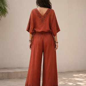 May include: Rust-colored two-piece outfit featuring a V-neck top with embroidered detailing and wide-leg pants. The top has short sleeves and drawstring ties at the waist. The pants have an embroidered design at the hem. The outfit is made of a lightweight fabric.