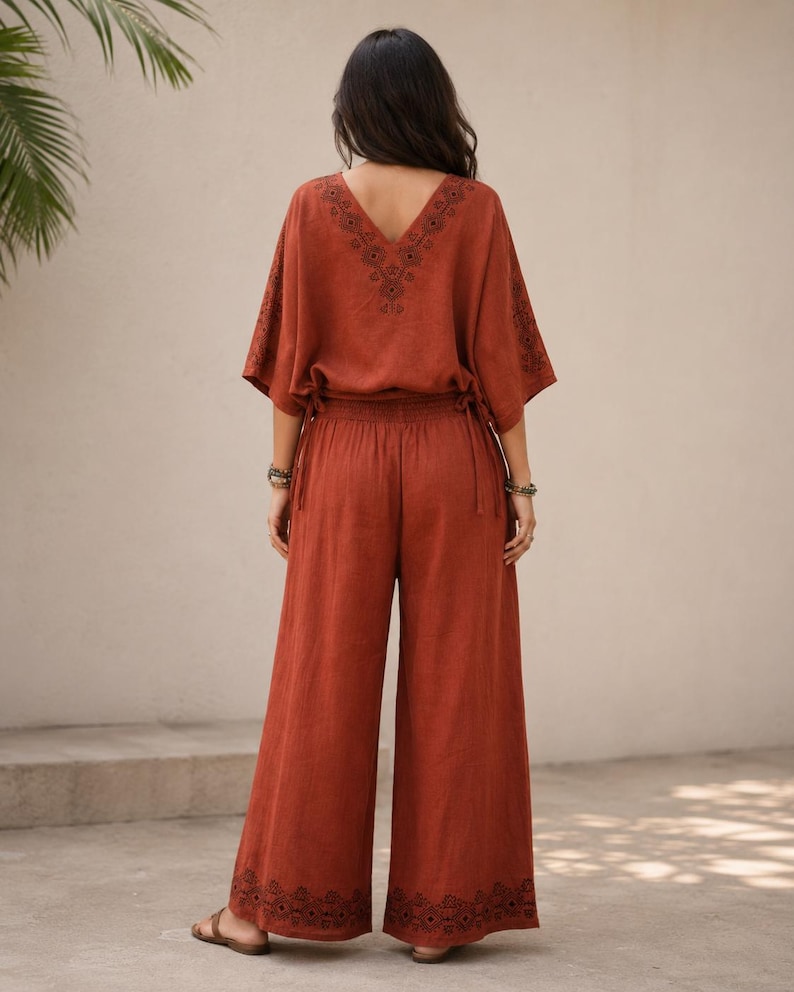 May include: Rust-colored two-piece outfit featuring a V-neck top with embroidered detailing and wide-leg pants. The top has short sleeves and drawstring ties at the waist. The pants have an embroidered design at the hem. The outfit is made of a lightweight fabric.