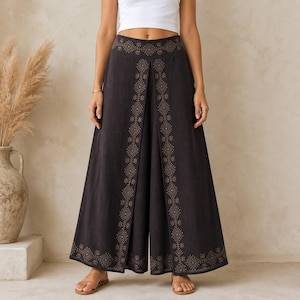 May include: Wide-leg, dark charcoal pants with a decorative gold geometric pattern along the front and hem. The pants have a high waist and a split front design. The model is wearing a white top and sandals.