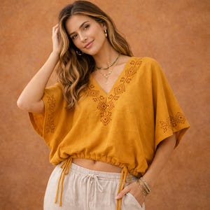 May include: A mustard yellow short-sleeve blouse featuring a V-neckline with dark brown geometric embroidery, wide sleeves, and a cinched hemline with adjustable drawstring ties at the sides, paired with light beige drawstring pants.