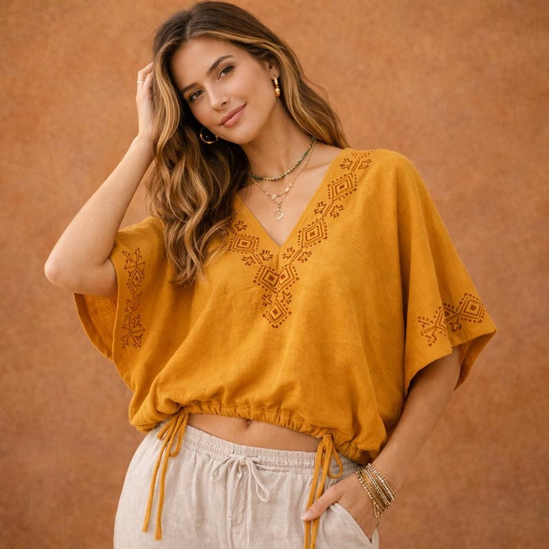May include: A mustard yellow short-sleeve blouse featuring a V-neckline with dark brown geometric embroidery, wide sleeves, and a cinched hemline with adjustable drawstring ties at the sides, paired with light beige drawstring pants.