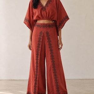 May include: A rust-colored two-piece outfit featuring a cropped top with a V-neck and wide-leg pants. The top and pants are adorned with black embroidered patterns. The outfit is made of a lightweight fabric, perfect for warm weather.