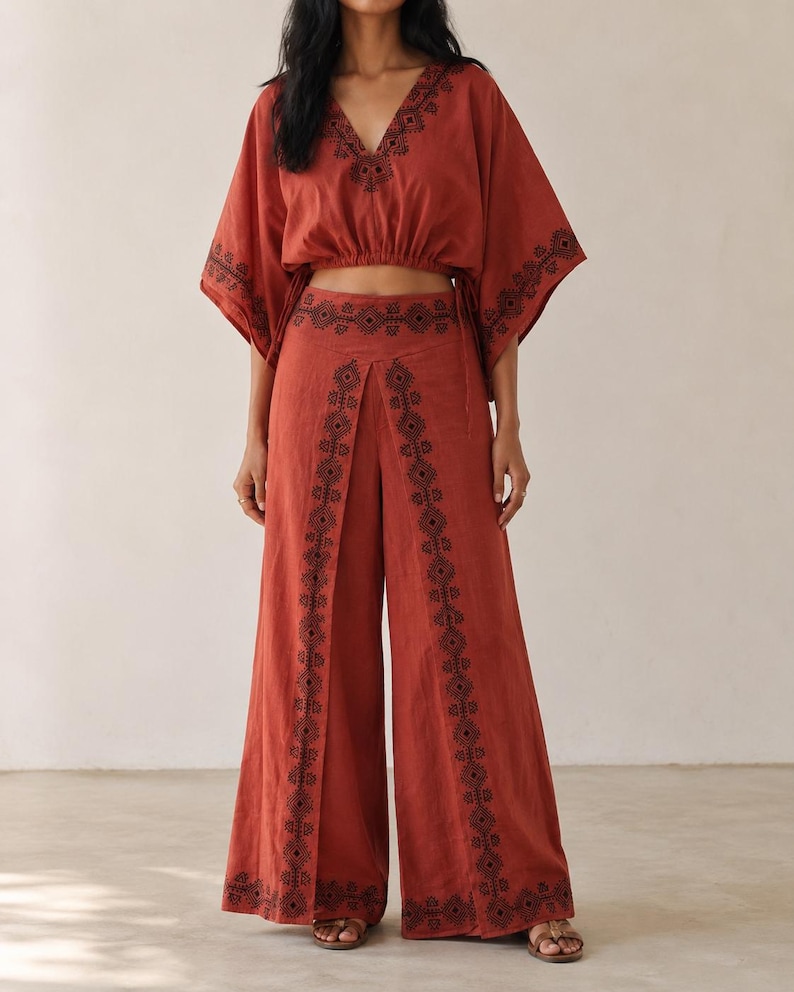 May include: A rust-colored two-piece outfit featuring a cropped top with a V-neck and wide-leg pants. The top and pants are adorned with black embroidered patterns. The outfit is made of a lightweight fabric, perfect for warm weather.