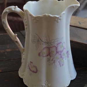 May include: A white ceramic pitcher with a scalloped rim and base, featuring a delicate floral pattern in shades of purple and green. The pitcher has a curved handle with embossed detailing and a fluted body, showcasing a soft, painted aesthetic.
