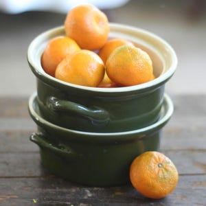 May include: Two dark green ceramic bowls with white rims are stacked, filled with bright orange tangerines. One tangerine rests outside the bowls on a wooden surface. The bowls have small handles on each side.