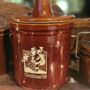 May include: A brown ceramic crock with a lid, featuring a Santa Claus graphic and the text "Christmas '88". The crock has a handle and a lid. The overall theme is vintage Christmas decor.
