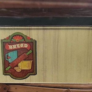 May include: A rectangular bread box with a black lid and a light yellow body. The front features a decorative sign with the word "BREAD" and illustrations of baking tools. The box is made of metal and has a vintage aesthetic.