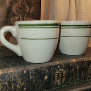 May include: Two white ceramic coffee cups with green horizontal stripes. The cups have a rounded shape with a handle on the side. The cups are sitting on a dark surface.