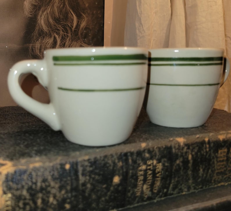 May include: Two white ceramic coffee cups with green horizontal stripes. The cups have a rounded shape with a handle on the side. The cups are sitting on a dark surface.