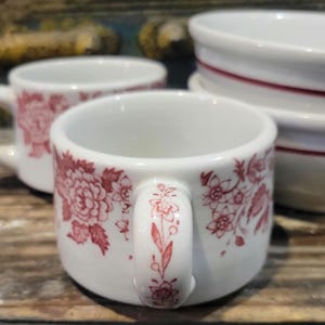 May include: White ceramic cups with a red floral pattern. The cups have a handle with a floral design. The cups are stacked on top of each other. The cups are on a wooden surface.