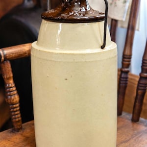 May include: A cylindrical stoneware crock with a light cream-colored body and a dark brown glazed lid. The lid features a metal wire bail handle attached to the sides. The surface of the crock is smooth with subtle variations in the off-white finish.