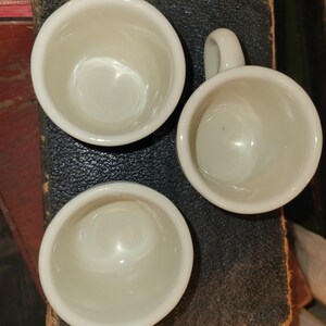 May include: Three small, white ceramic cups with handles are arranged on a dark, textured surface. The cups are round with a smooth, glossy finish, and the handles are small and curved. The cups are empty and appear to be clean.
