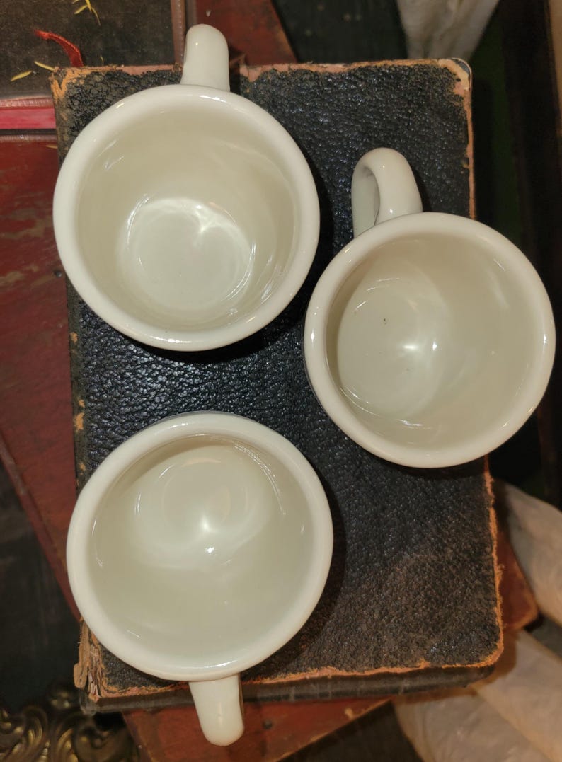 May include: Three small, white ceramic cups with handles are arranged on a dark, textured surface. The cups are round with a smooth, glossy finish, and the handles are small and curved. The cups are empty and appear to be clean.