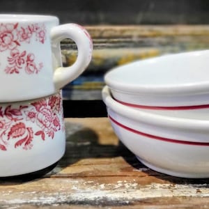 May include: Two white ceramic mugs with red floral patterns stacked on top of each other. Beside them are three white bowls with a red stripe around the rim. The items are on a wooden surface.