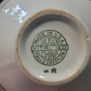 May include: A close-up of the bottom of a white ceramic piece. The text "MADE IN U.S.A. JACKSON VITRIFIED CHINA FALLS CREEK, PA" is printed in green within a circular emblem. The number "A6" and the letter "R" are also visible.
