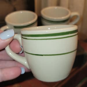 May include: Cream-colored ceramic espresso cups with green stripes. The cups have small handles and a classic diner style. Several cups are visible, with one held in a hand with light blue nail polish.
