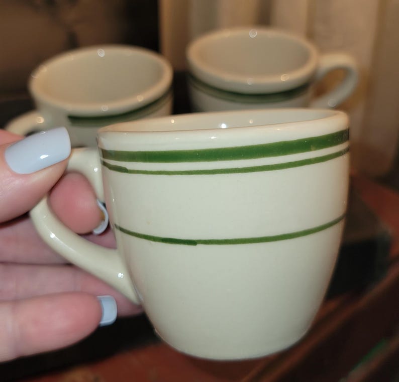 May include: Cream-colored ceramic espresso cups with green stripes. The cups have small handles and a classic diner style. Several cups are visible, with one held in a hand with light blue nail polish.