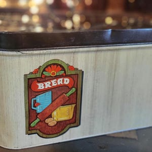 May include: Vintage bread box with a brown lid and a cream-colored body. The box features a decorative label with the word "BREAD" and illustrations of a rolling pin, measuring cup, and loaf of bread. The box has a rectangular shape.