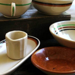 May include: A collection of ceramic tableware including a cream-colored cup with a green stripe, a bowl with brown and black bands, a small cream-colored pitcher on an oval tray, a bowl with green and brown stripes, and a small solid brown bowl.