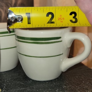 May include: White ceramic coffee cup with two horizontal green stripes. The cup is approximately 2.5 inches in diameter and has a curved handle. The cup is being measured with a yellow tape measure.
