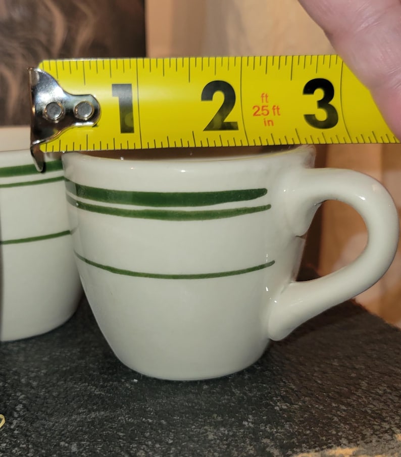 May include: White ceramic coffee cup with two horizontal green stripes. The cup is approximately 2.5 inches in diameter and has a curved handle. The cup is being measured with a yellow tape measure.