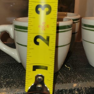 May include: White ceramic espresso cups with a green stripe. The cups have a handle and are approximately 2.5 inches tall. The cups are on a dark surface.