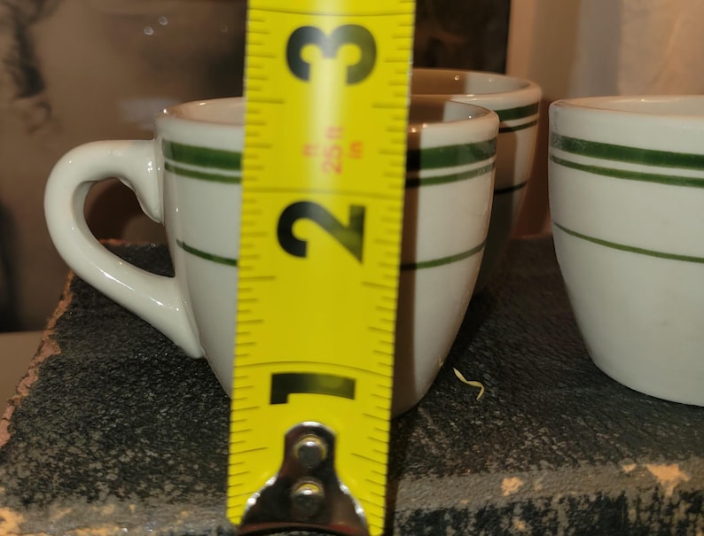 May include: White ceramic espresso cups with a green stripe. The cups have a handle and are approximately 2.5 inches tall. The cups are on a dark surface.