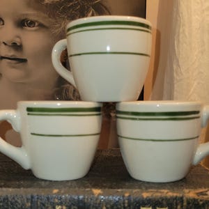 May include: Three white ceramic mugs with a green stripe design. Each mug has a curved handle and a rounded body. The mugs are stacked on top of each other. The background includes a vintage photograph and a curtain.