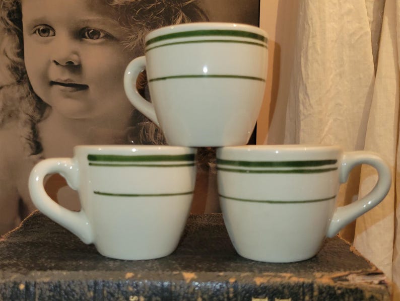 May include: Three white ceramic mugs with a green stripe design. Each mug has a curved handle and a rounded body. The mugs are stacked on top of each other. The background includes a vintage photograph and a curtain.