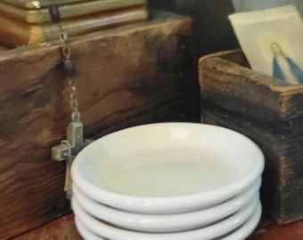 May include: A stack of four small, round, white ceramic saucers with smooth, curved edges. The saucers are plain, featuring a glossy finish and a shallow, concave center, designed for holding small items or serving individual portions.