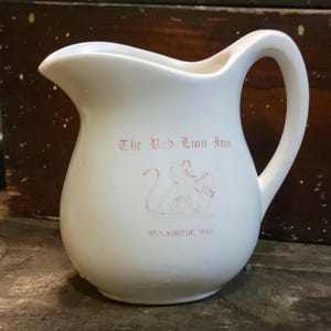 May include: A cream-colored ceramic pitcher featuring a curved handle and a pouring spout. The front displays the red text The Red Lion Inn, a graphic of a lion holding a shield with the year 1773, and the text STOCKBRIDGE, MASS. below it.