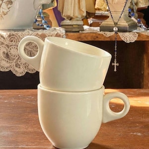 May include: Two stacked cream-colored ceramic cups with smooth, rounded bodies and distinct circular handles. The top cup is angled slightly, while the bottom cup sits upright, both featuring a simple, solid-colored finish and a clean, minimalist design.