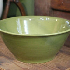 May include: A large, round, green ceramic bowl with a glossy finish. The bowl has a wide rim and a gently sloping side. The bowl is sitting on a wooden surface. The bowl is empty.