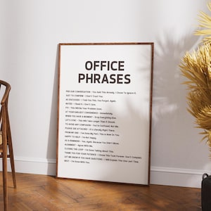 May include: A framed print with the words "OFFICE PHRASES" in bold black letters at the top. Below, a list of phrases in smaller black text. The frame is brown, and the print is displayed against a white wall.