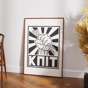 May include: A framed black and white print featuring a clenched fist holding knitting needles. The design includes a sunburst pattern and the word "KNIT" in bold letters, perfect for a craft room or as a gift for a knitter.